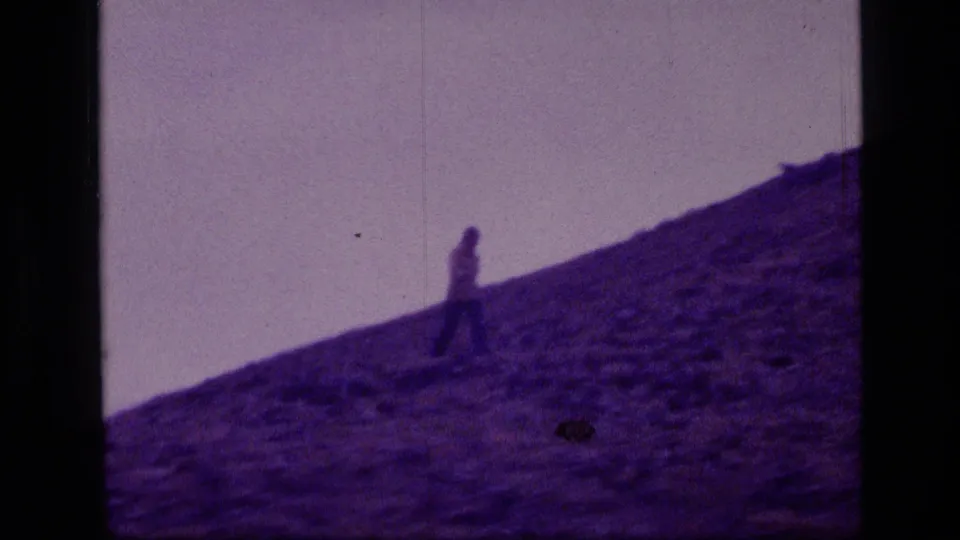 A lone walker moves along a slanted rocky slope in Scapegoat Wilderness, Montana, in purple-tinted 8mm home-movie footage.
