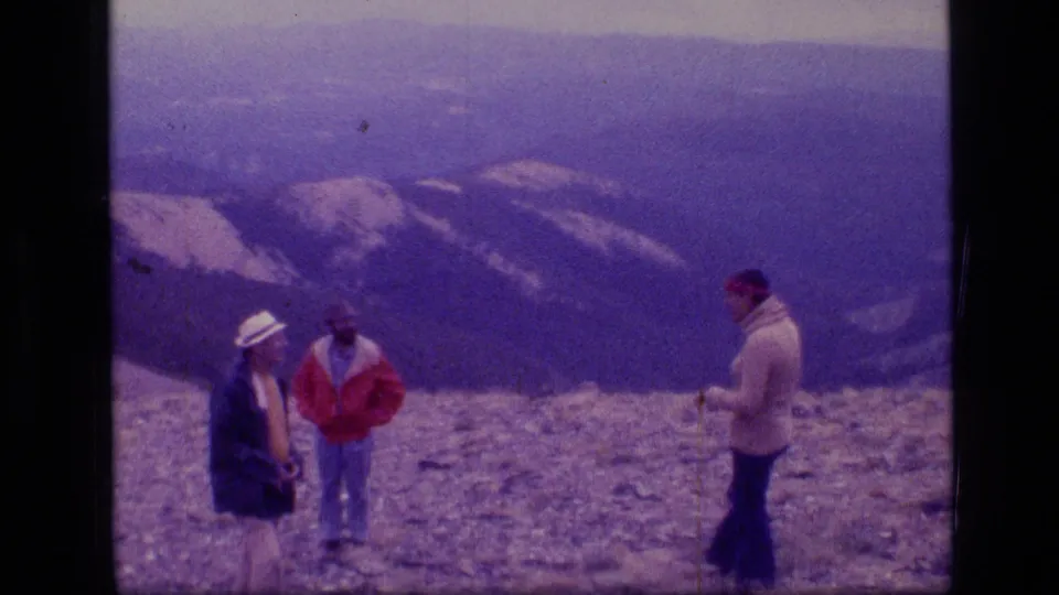 Three adults gather on a rocky summit overlook in 1973 Scapegoat Wilderness footage, with hazy Montana mountain ridges behind them.