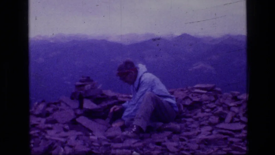 A hiker rests on a rocky Scapegoat Wilderness summit in 1973, with layered mountain ridges behind him.