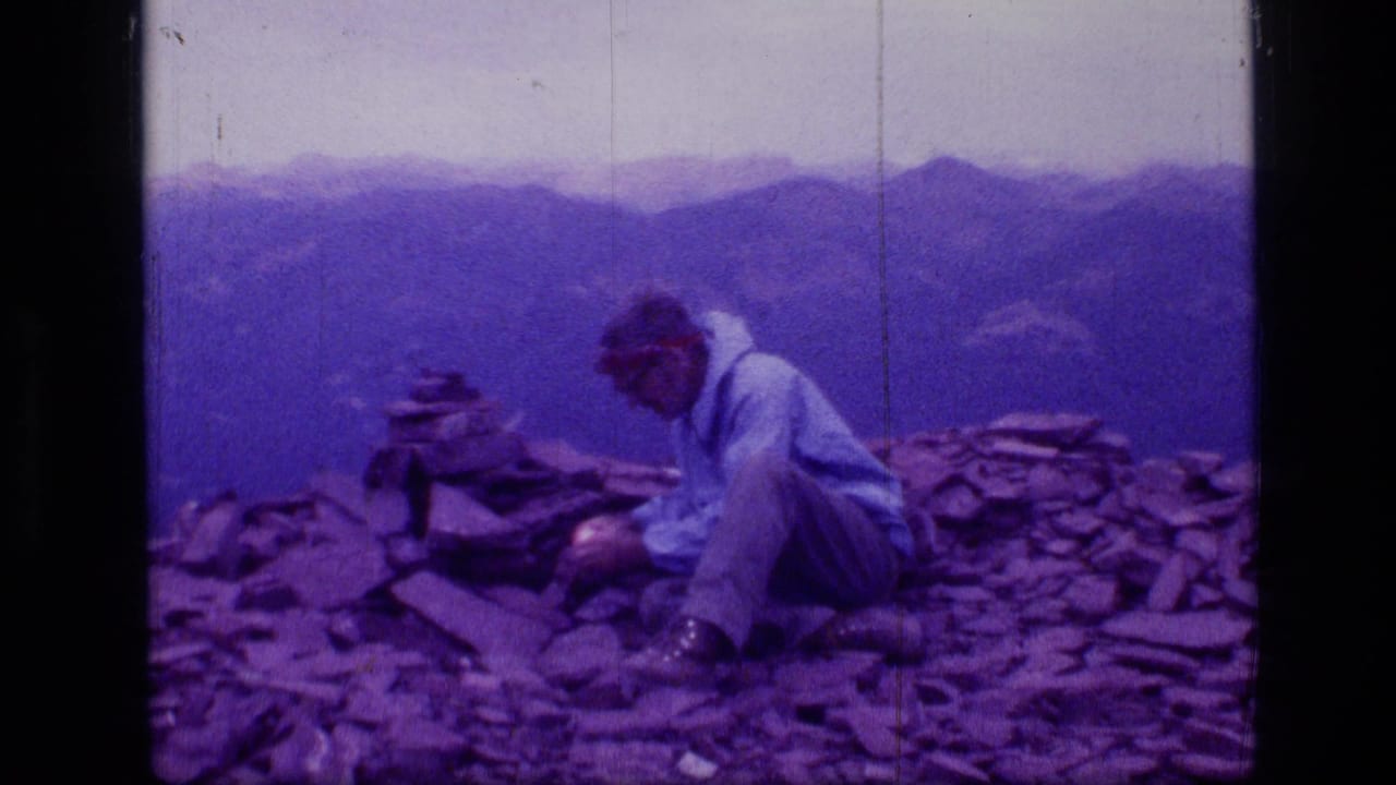 Man in a light jacket and hat sits on a rocky summit beside a small cairn with mountains behind him.