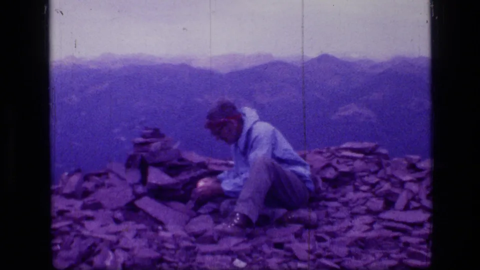 1973 Scapegoat Wilderness 8mm frame showing a seated man near a small cairn on a rocky mountain peak with distant ridges beyond.