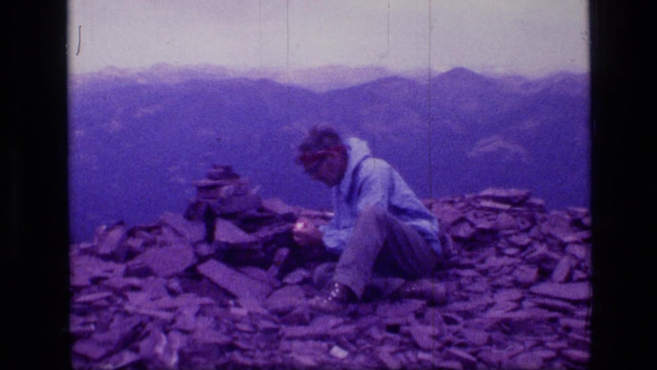 1973 Montana wilderness frame with a man seated on a rocky peak, handling something near a cairn against a hazy mountain backdrop.