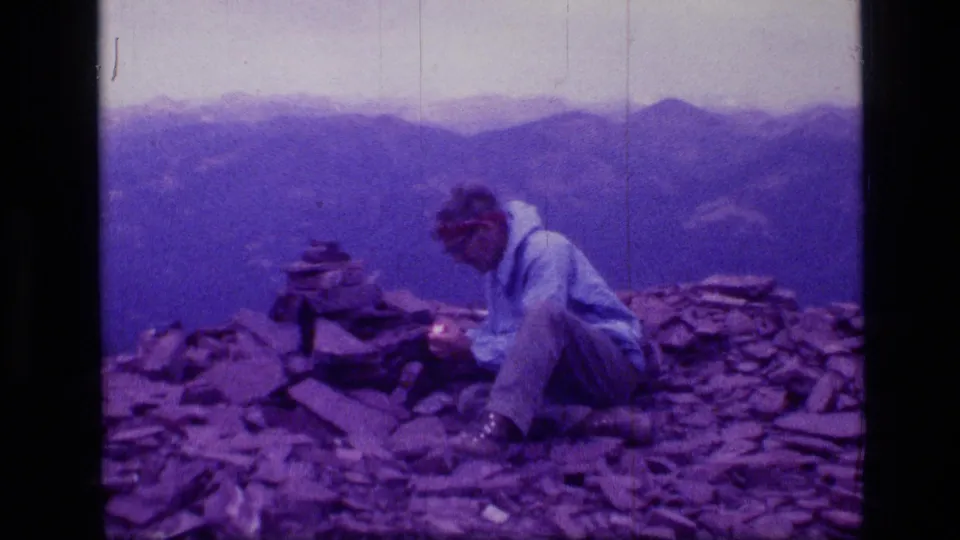 1973 Montana wilderness frame with a man seated on a rocky peak, handling something near a cairn against a hazy mountain backdrop.