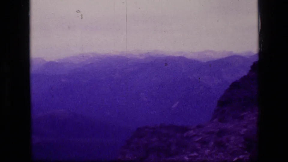 1973 Montana wilderness frame looking across layered mountain ridges from a rocky high point, rendered with aged 8mm color shift.