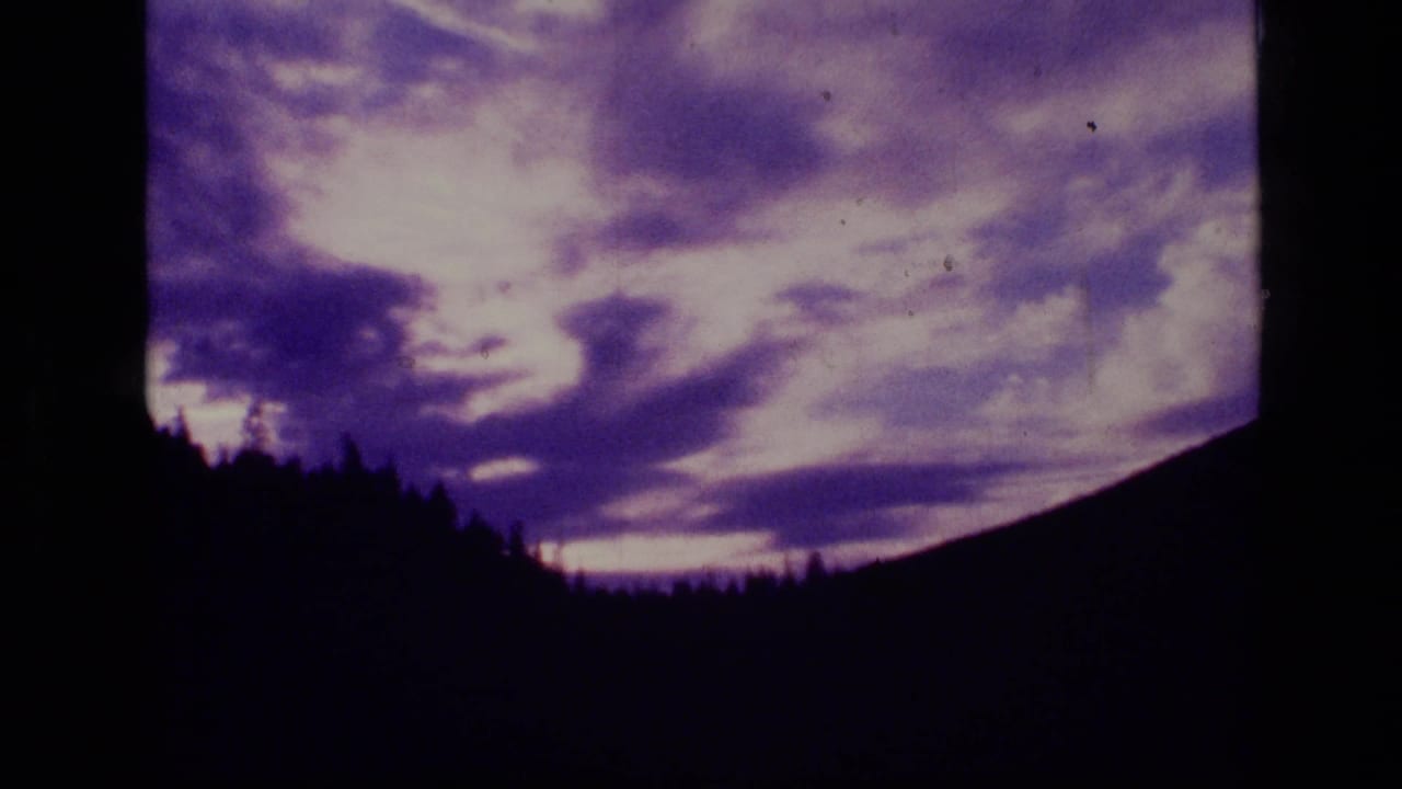 1973 Montana 8mm frame with bright purple sunset clouds behind a black treeline and mountain ridge.