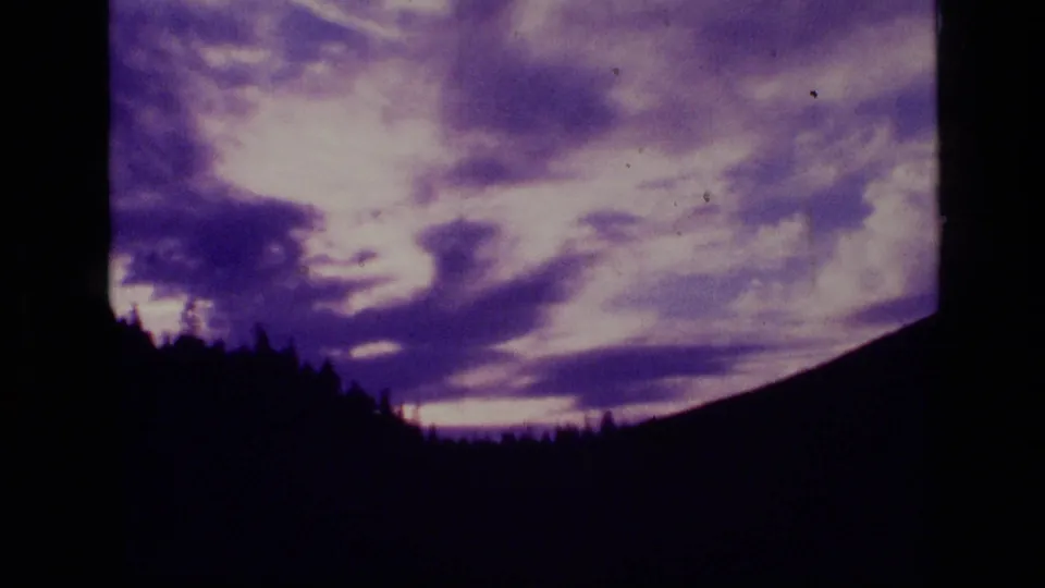 1973 Montana 8mm frame with bright purple sunset clouds behind a black treeline and mountain ridge.