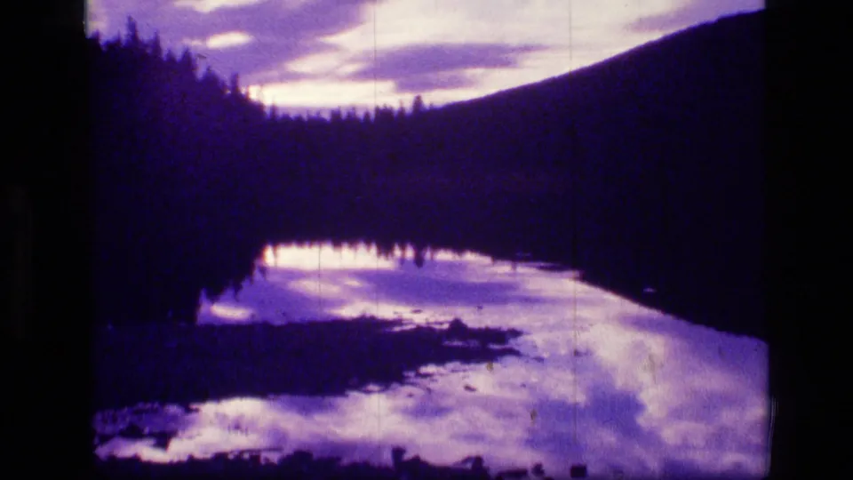1973 Montana wilderness 8mm frame with a shadowed lake reflecting violet clouds below forested hills and a mountain slope.