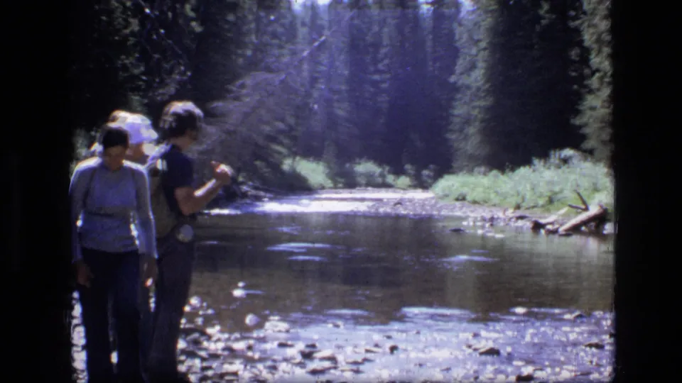 Two people pause along a rocky riverbank in Scapegoat Wilderness, Montana, with forest continuing into the background.