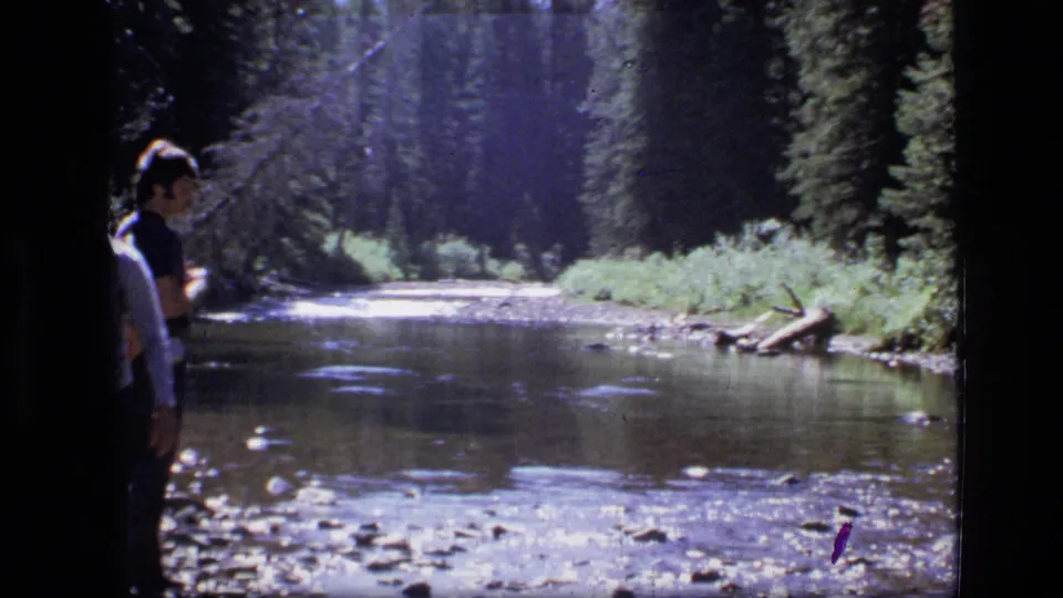 In 1973 Scapegoat Wilderness footage, two visitors stand near a shallow rocky river bordered by dense evergreens.