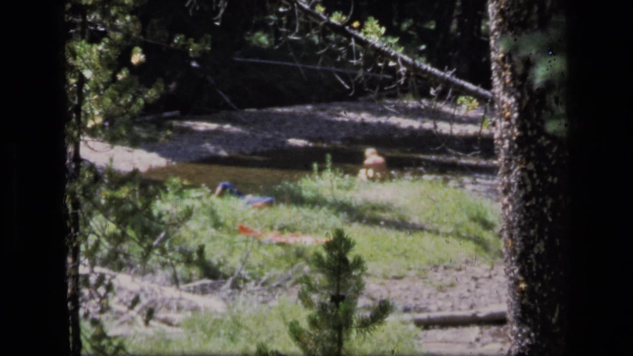 1973 8mm Montana wilderness view of a man resting beside a shallow creek, seen through trees with fabric items nearby.