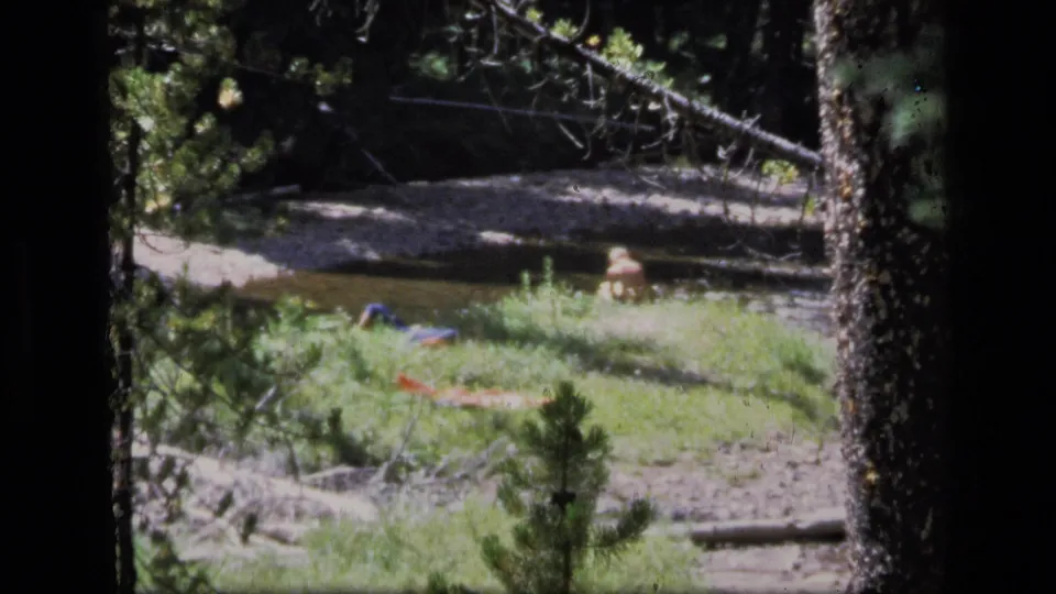 1973 8mm Montana wilderness view of a man resting beside a shallow creek, seen through trees with fabric items nearby.