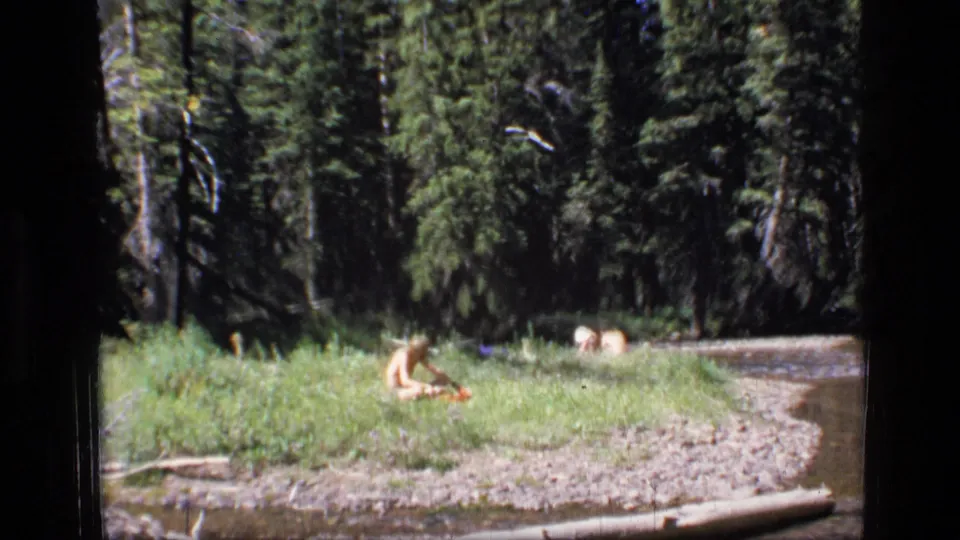8mm home-movie frame from the 1973 Montana clip showing a child in grass near a gravel bend and shaded evergreen woods.