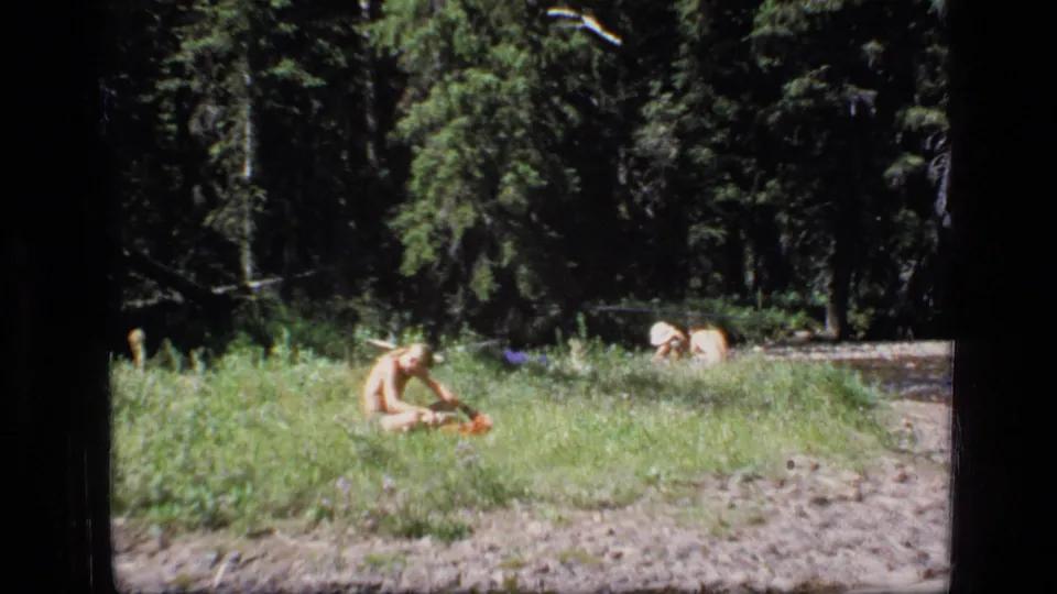 Final 1973 Scapegoat Wilderness frame showing a child seated by a slow river with fallen wood, gravel shore, and tall evergreens.