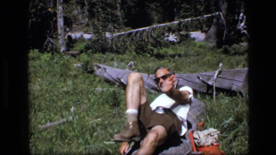Adult man in sunglasses, white shirt, brown shorts, socks, and boots resting in a grassy Scapegoat Wilderness clearing.