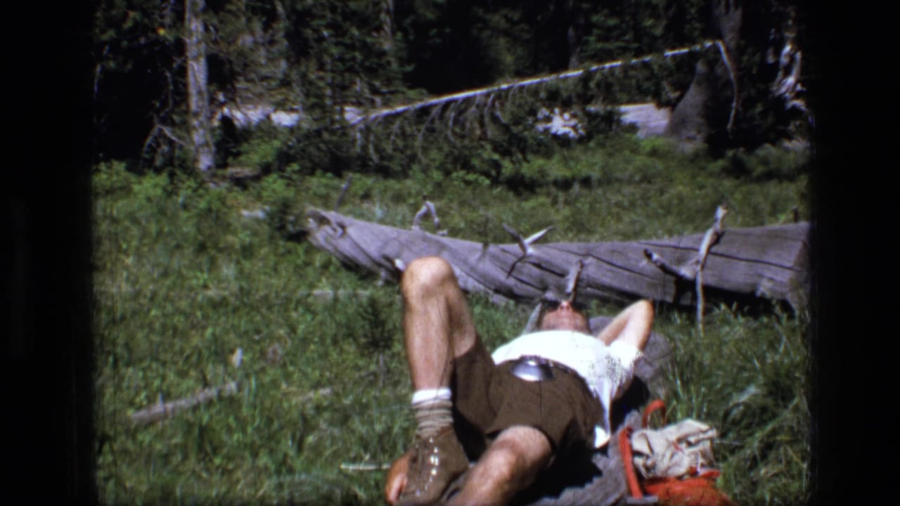 1973 Montana wilderness home-movie frame showing a male camper resting on his back near a fallen log, with grass, trees, boots, and a pack visible.