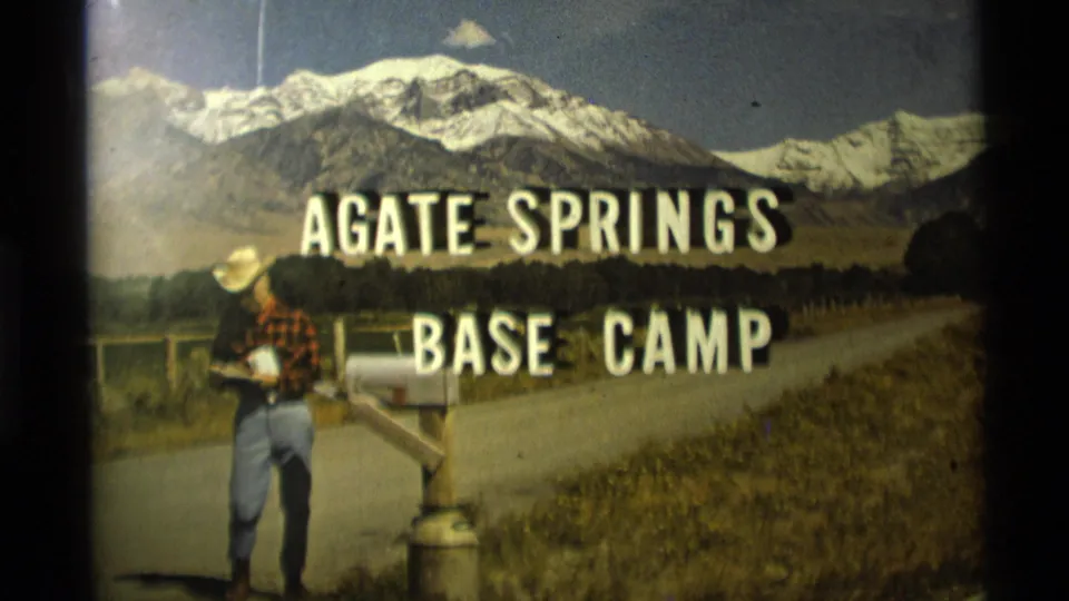 1975 Montana home-movie frame with Agate Springs Base Camp title text, a man by a mailbox, a rural road and snow-covered mountains.