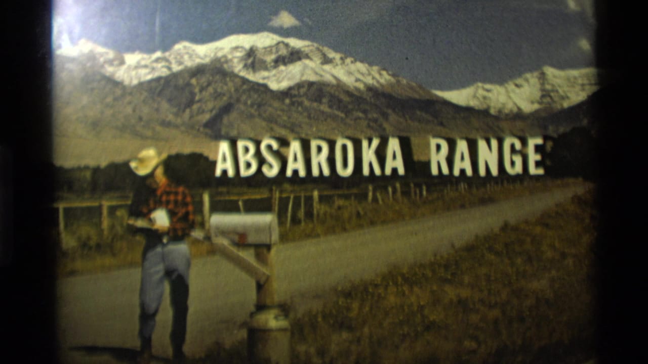 Man stands by a mailbox beneath large Absaroka Range sign text and snowy mountains.