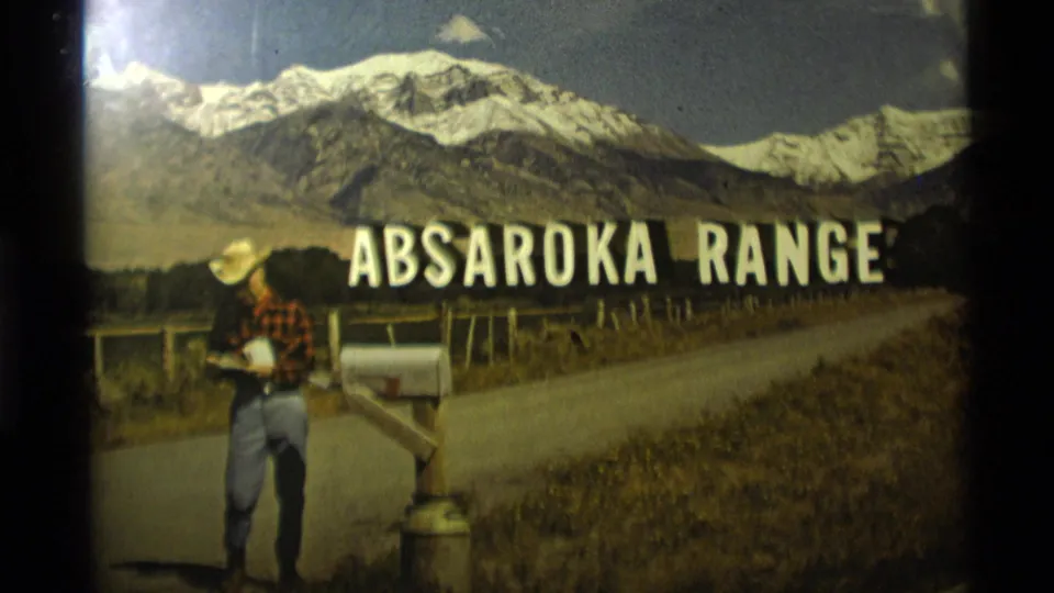 1975 Montana home-movie frame with a cowboy-hatted man by a mailbox and Absaroka Range sign, backed by snow-capped peaks.