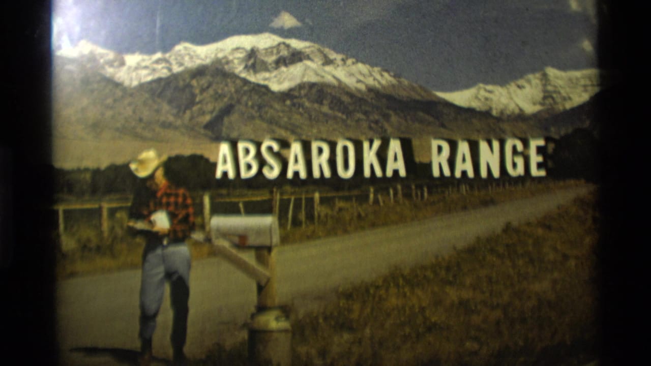 1975 Montana home-movie frame showing a man in a cowboy hat beside a roadside mailbox below the Absaroka Range title and snow-capped mountains.
