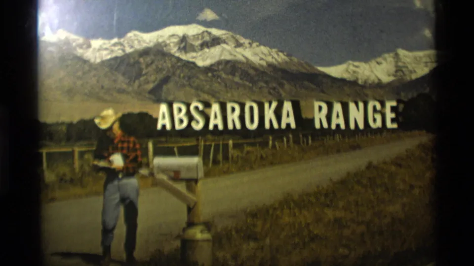 1975 Montana home-movie frame showing a man in a cowboy hat beside a roadside mailbox below the Absaroka Range title and snow-capped mountains.