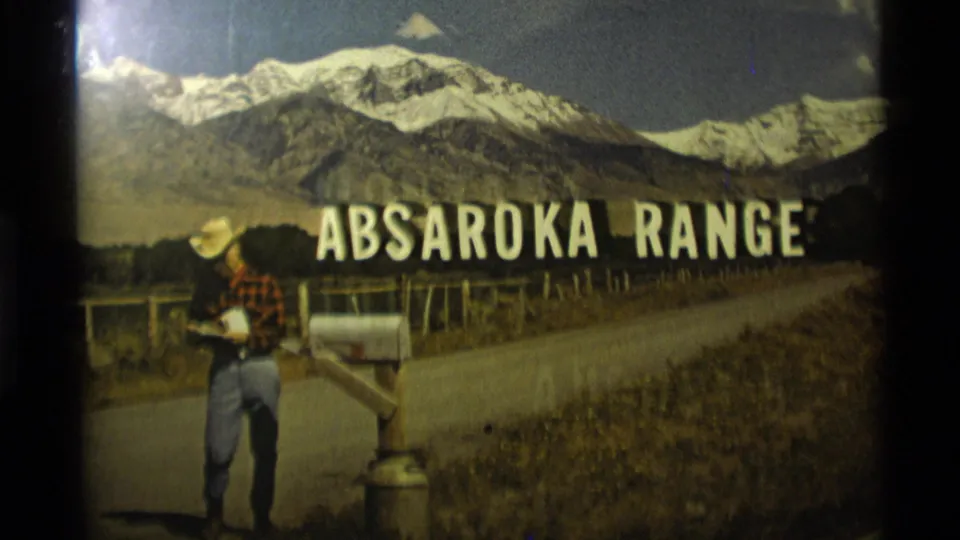 Small-gauge Montana frame showing the same roadside mailbox, western-dressed man, fence, road, and Absaroka Range title against snow-covered peaks.