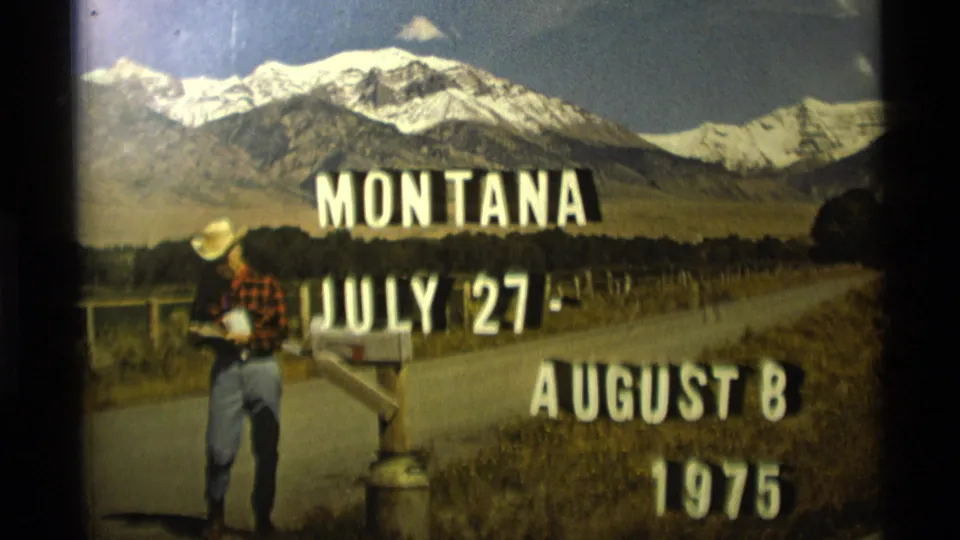 Home-movie frame from Montana showing July-August 1975 title text over a rural road, mailbox, and mountain backdrop.