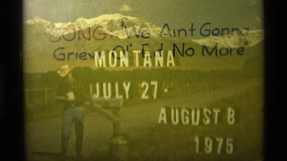 Faded 1975 Montana 8mm frame showing trip-title text beneath a handwritten note about a song, with the roadside figure still faintly visible.