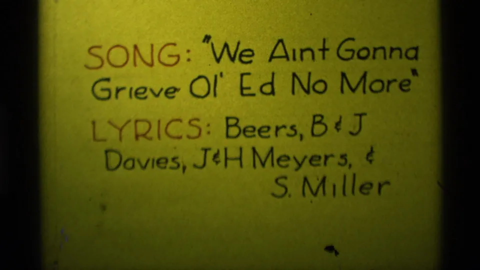 Close 1975 8mm view of a yellow handwritten card naming the song and listing lyricist credits in marker.