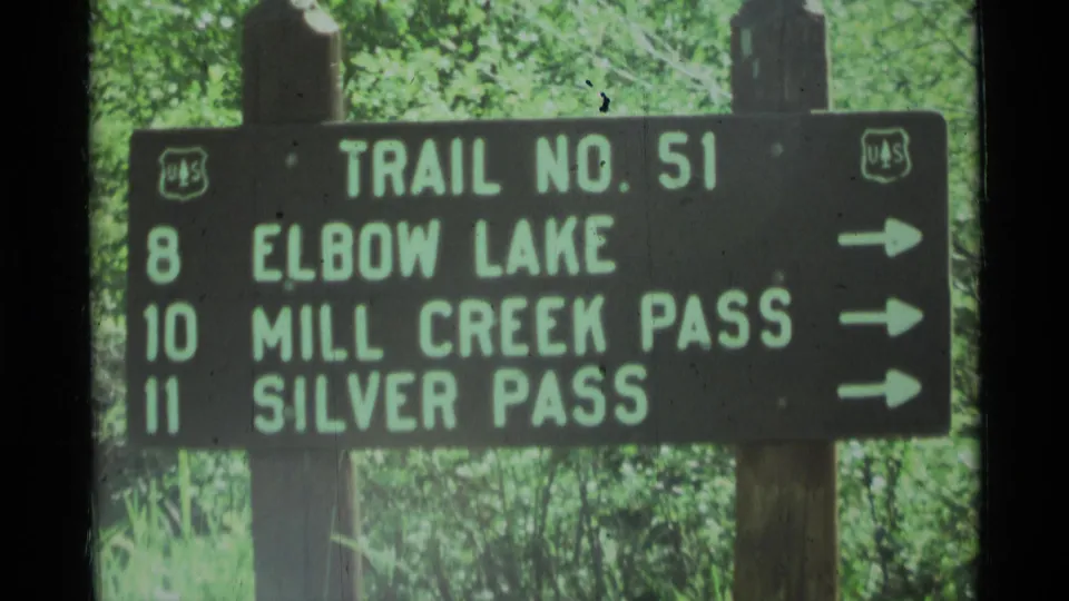 1975 Montana 8mm frame holds on the Trail No. 51 sign, with destination text, arrows, wood posts, and forest background visible.