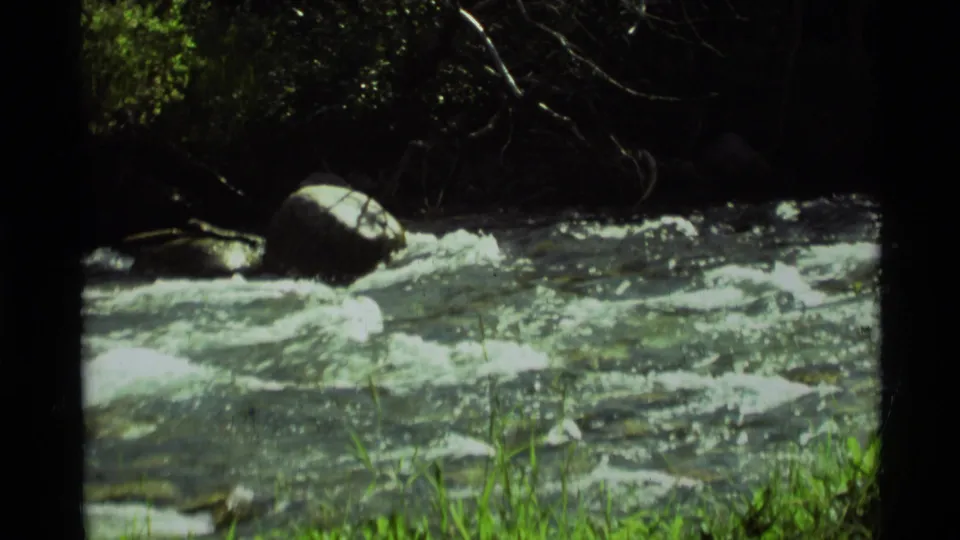 1975 8mm Montana frame showing a bright mountain creek rushing over rocks with shaded trees and green grass along the bank.