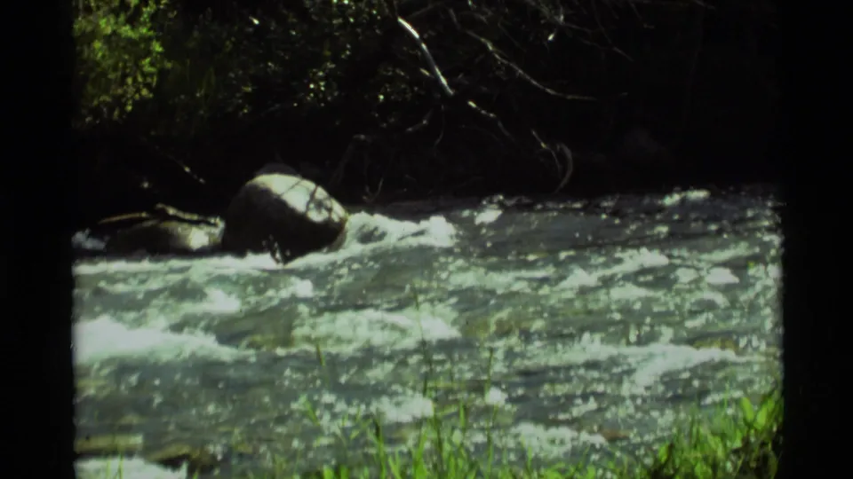 1975 8mm Montana frame showing fast creek water, rocks, and dark tree cover near a grassy bank.