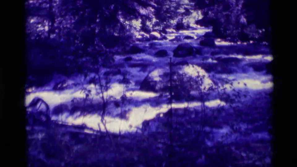 Agate Springs home-movie frame showing bright rapids winding past rocks under dense, purple-shifted foliage.