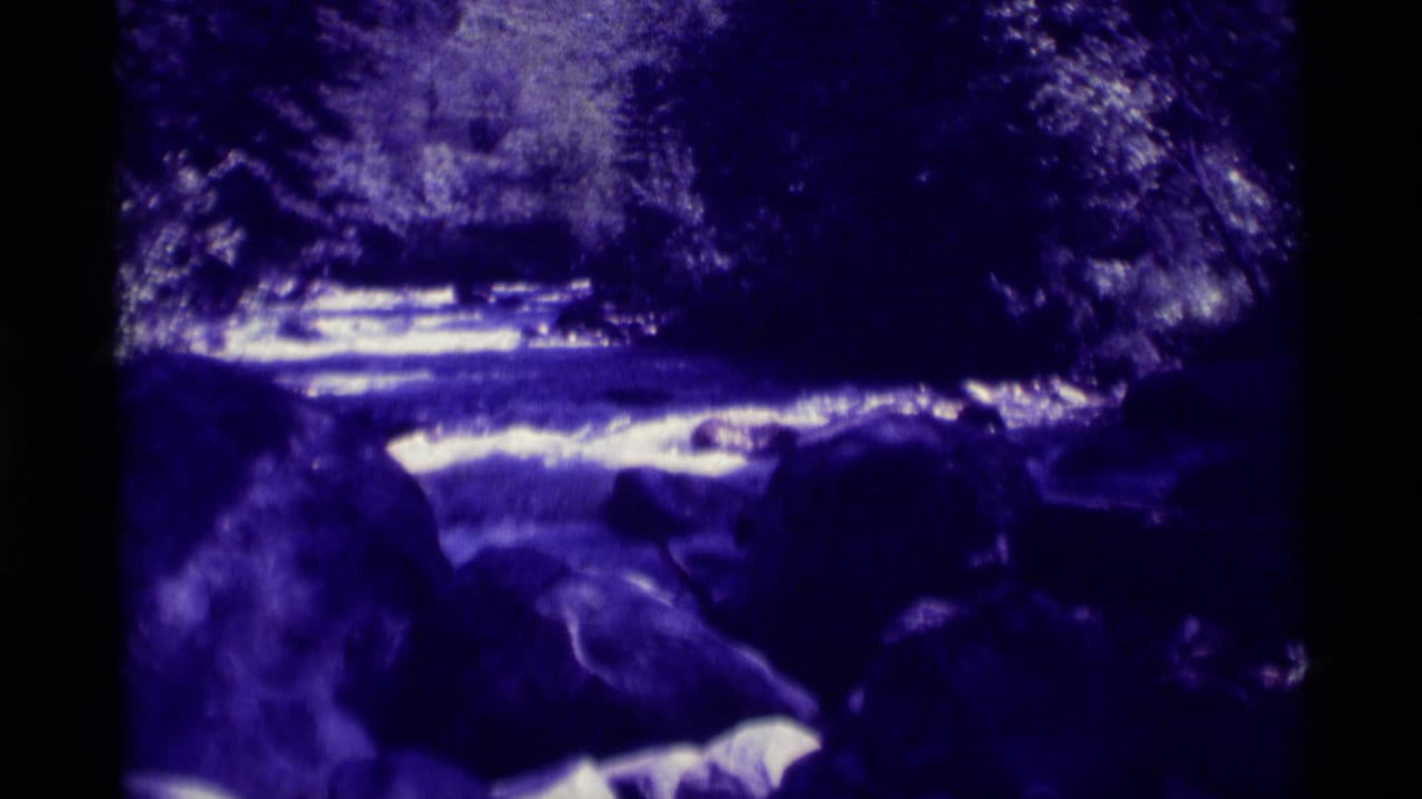 1975 8mm view of water moving over large rocks at Agate Springs, Montana, framed by trees and deep shadows.