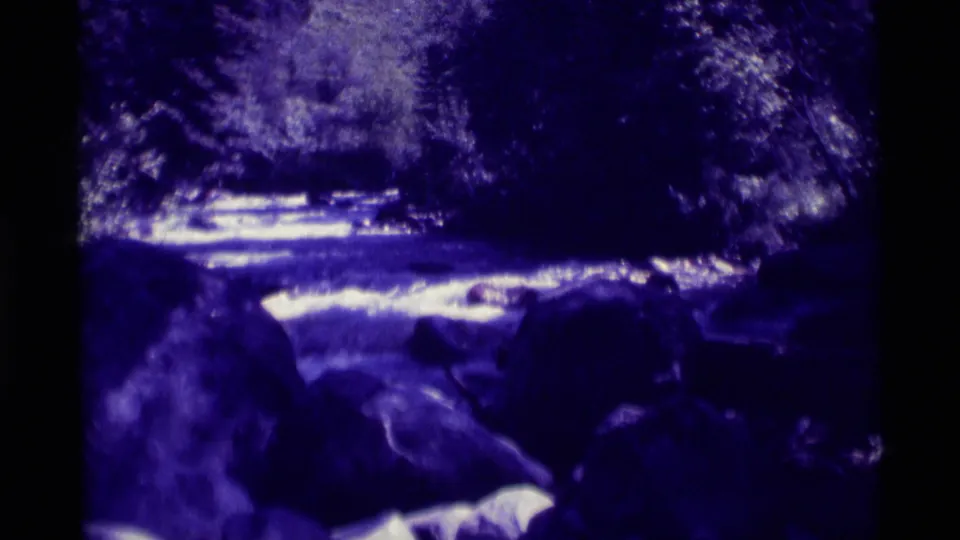 1975 8mm view of water moving over large rocks at Agate Springs, Montana, framed by trees and deep shadows.
