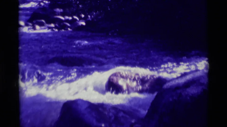 A blue-tinted 1975 8mm frame from Agate Springs, Montana shows clean-looking river water rushing past rocks and shadowed trees.