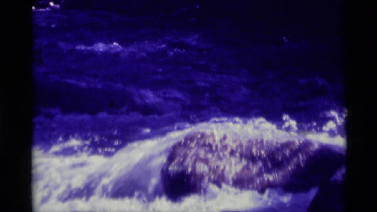 1975 8mm Montana footage showing rushing mountain river water around rocks with dark frame edges and purple-blue color shift.