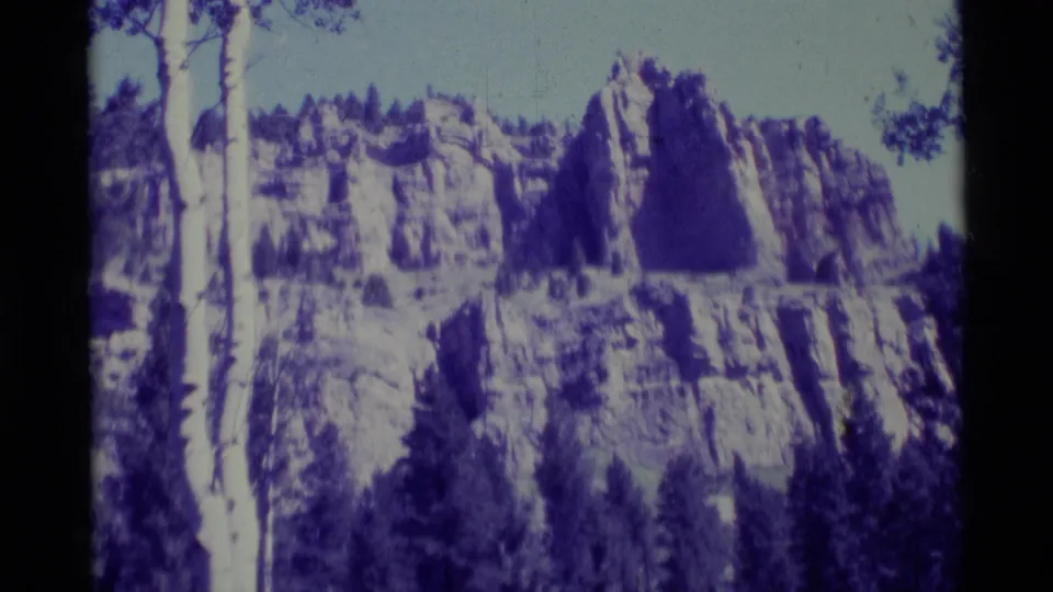 1975 8mm home-movie frame of a rocky mountainside in Agate Springs, Montana, with conifers and a strong blue-purple film cast.