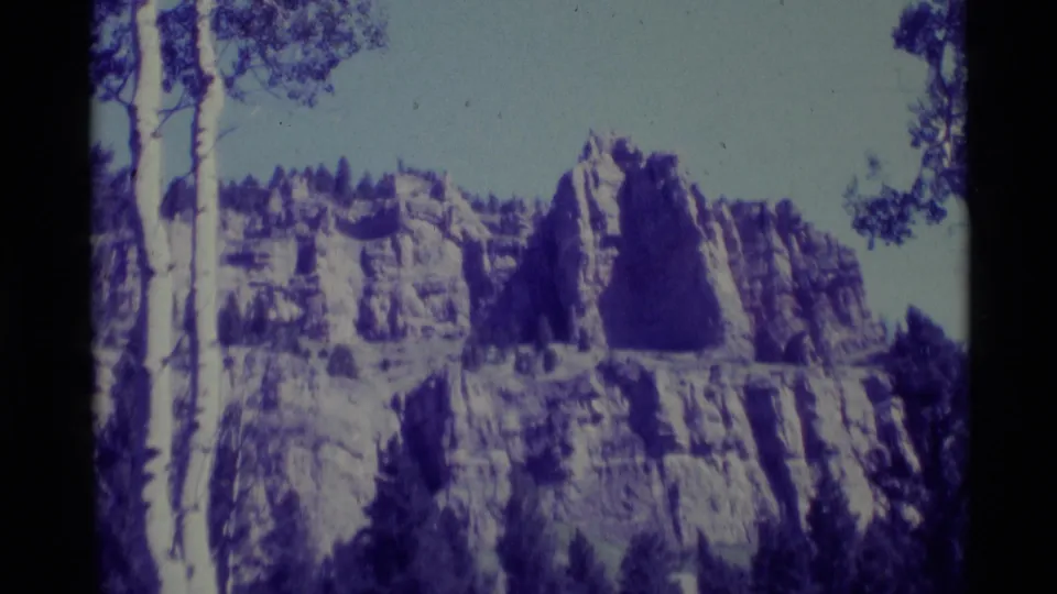 1975 8mm view of a rocky Montana mountainside near Agate Springs, showing cliffs, conifers, pale sky, and home-movie color shift.