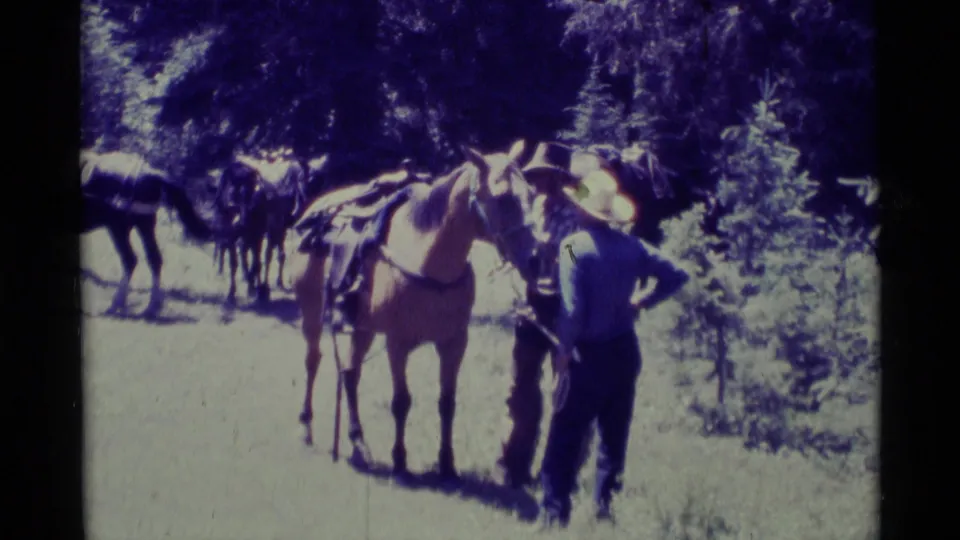 1975 Agate Springs frame showing two hat-wearing men tending horses in a sunlit wooded clearing.