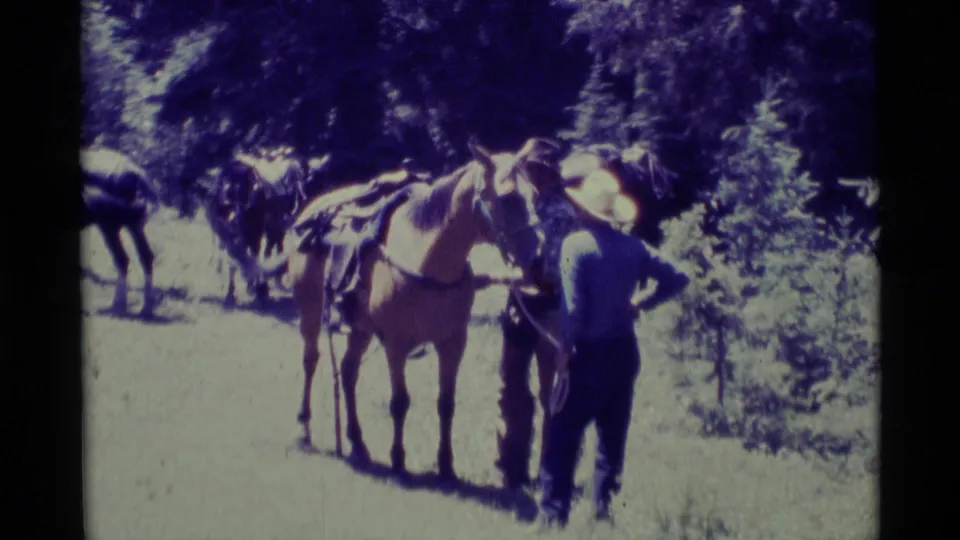 1975 Agate Springs, Montana home-movie frame showing an adult man in a cowboy hat beside a saddled horse with other horses behind him.