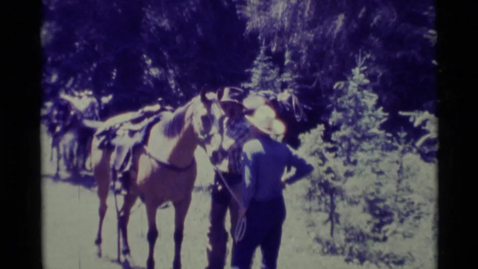 1975 Montana home-movie frame with a cowboy-hatted adult standing close to a saddled horse in a wooded outdoor setting.