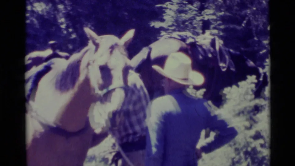 Close 1975 Agate Springs frame showing two adult men in cowboy hats near a saddled horse, with trees behind them.