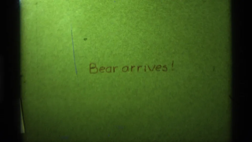 Green-tinted title card from the home movie, showing Bear arrives! in brown handwriting with visible scratches and edge shadow.