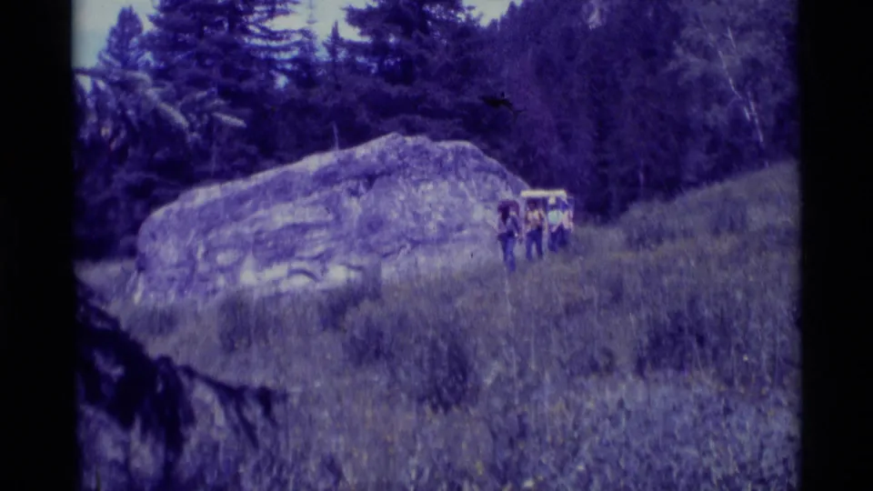 Three backpacked hikers in a purple-tinted 1975 8mm view of a grassy meadow at Agate Springs, Montana, with a large boulder and evergreens behind them.