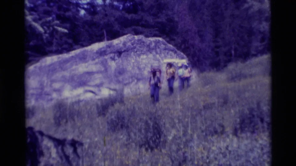 The 1975 Montana home-movie view shows four backpackers moving down the slope past rock and trees.