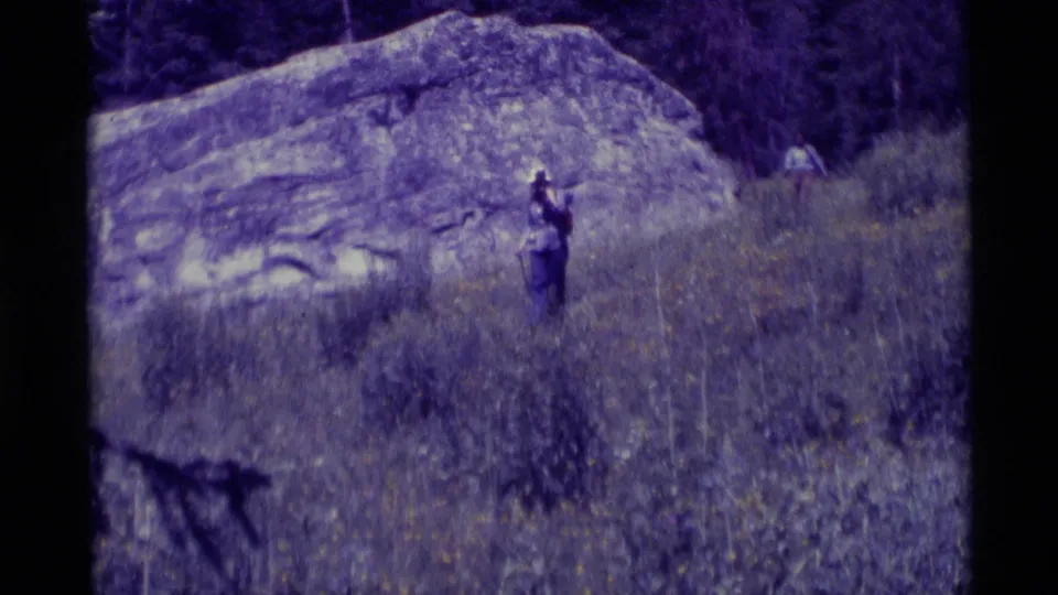 A hiker appears small in the landscape, walking through dry grass below a large boulder in 1975 Agate Springs footage.