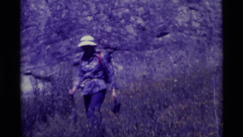 A backpacked hiker wearing a wide-brimmed hat walks through dry grass near rocky outcrops in the Montana home-movie scene.