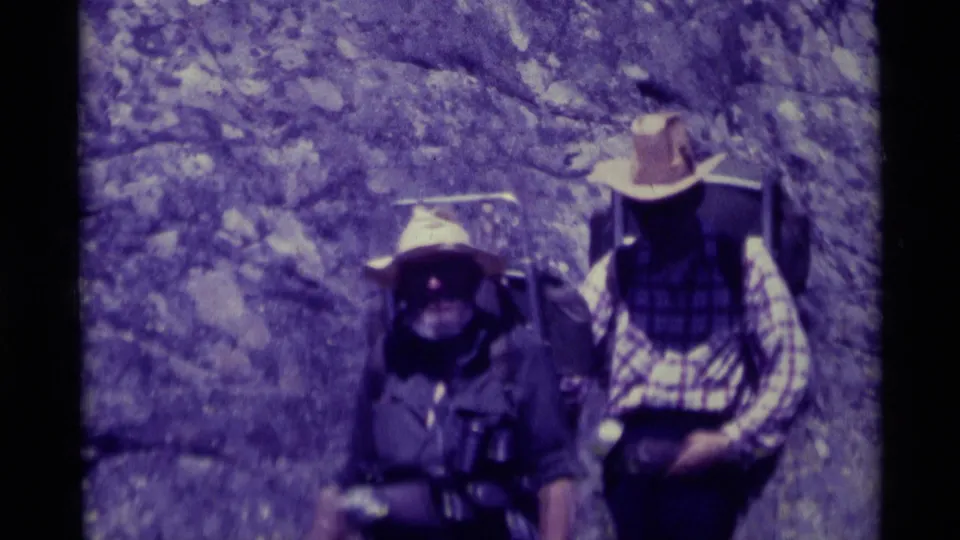 A closer 8mm view shows two 1970s hikers in hats, one in plaid, carrying backpacks near a rock formation in Montana.