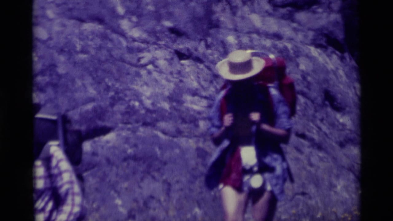 A 1975 8mm frame from Agate Springs, Montana, showing a hiker with a pack and wide-brimmed hat against a rocky hillside.