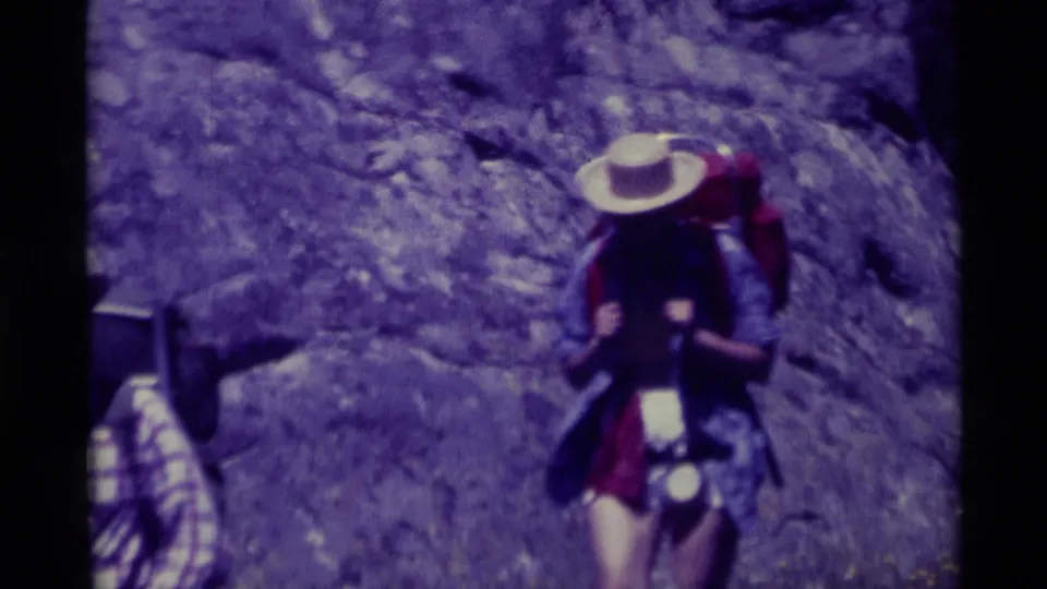 A 1975 8mm frame from Agate Springs, Montana, showing a hiker with a pack and wide-brimmed hat against a rocky hillside.